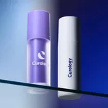Two Curology skincare bottles, one purple and one white, are displayed on a reflective glass surface with a gradient blue background.