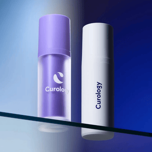 Two Curology skincare bottles, one purple and one white, are displayed on a reflective glass surface with a gradient blue background.
