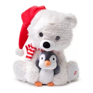 A plush polar bear wearing a red Santa hat and striped scarf is holding a small penguin. The bear's paw features a music note, suggesting it might be musical.