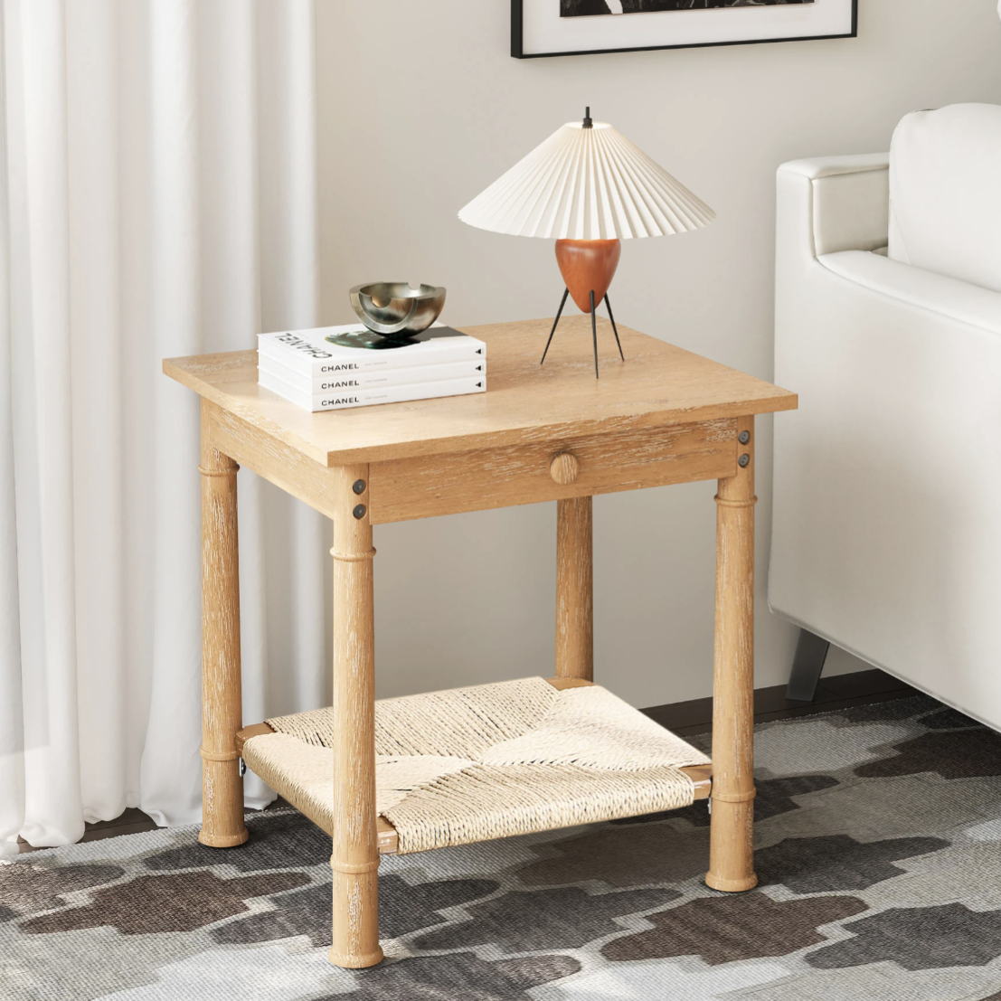 A wooden side table with a woven shelf holds a table lamp with a pleated shade and metallic legs, a decorative bowl, and stacked books, placed beside a white sofa and a patterned rug.
