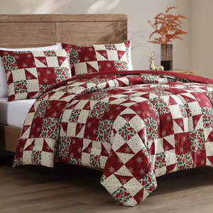 A quilt set with a red and cream patchwork design featuring holiday motifs like holly and snowflakes, includes a comforter and matching pillow shams on a bed with a wooden headboard.