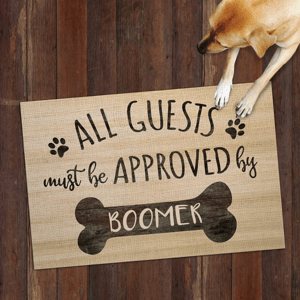 A doormat with the text \“All guests must be approved by Boomer\“ features paw prints and a bone design. A dog is seen resting its paws on the doormat.