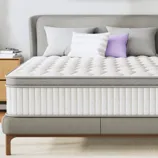 A queen-sized mattress with a quilted white and gray design is on a modern bed frame, surrounded by neutral decor including a lavender abstract painting, side tables with a lamp and clock, and decorative pillows.