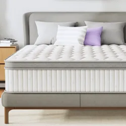 A queen-sized mattress with a quilted white and gray design is on a modern bed frame, surrounded by neutral decor including a lavender abstract painting, side tables with a lamp and clock, and decorative pillows.