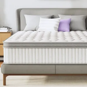 A queen-sized mattress with a quilted white and gray design is on a modern bed frame, surrounded by neutral decor including a lavender abstract painting, side tables with a lamp and clock, and decorative pillows.
