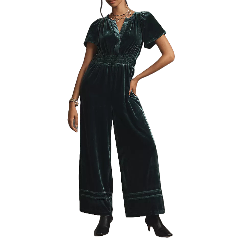 The Somerset Jumpsuit Velvet Edition features a deep green velvet fabric with short sleeves, a cinched waist, wide-leg pants, and a V-neckline, paired with black ankle boots.