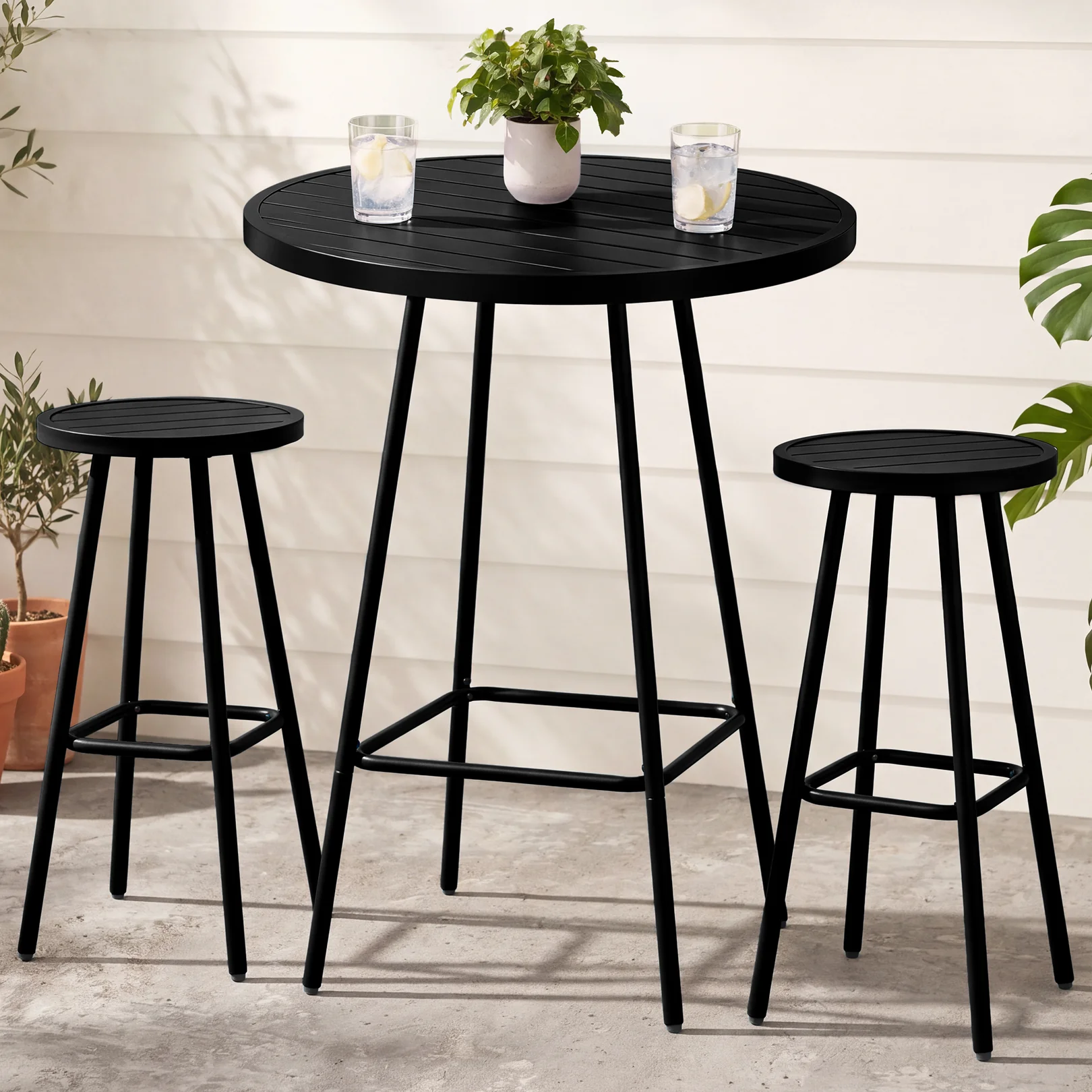A black round bar table with two matching stools, designed for outdoor use. The setup is decorated with potted plants and drinks, set on a concrete surface against a light paneled background.