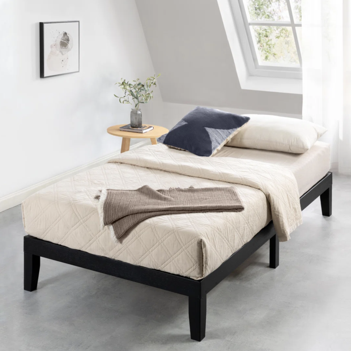 A minimalist bedroom setup with a twin bed featuring a beige quilted cover, navy and cream pillows, and a brown throw. A small round wooden side table holds a plant and a book, set against a window with natural light streaming in.
