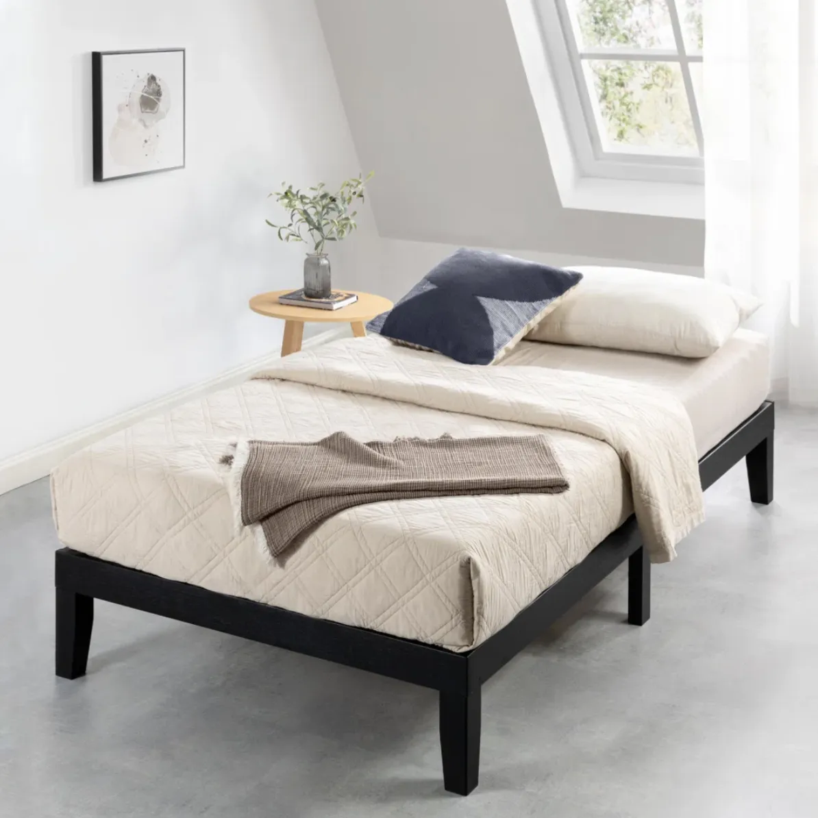 A minimalist bedroom setup with a twin bed featuring a beige quilted cover, navy and cream pillows, and a brown throw. A small round wooden side table holds a plant and a book, set against a window with natural light streaming in.