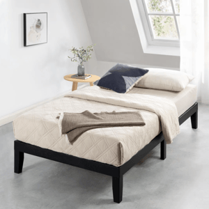 A minimalist bedroom setup with a twin bed featuring a beige quilted cover, navy and cream pillows, and a brown throw. A small round wooden side table holds a plant and a book, set against a window with natural light streaming in.