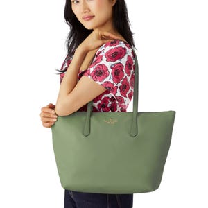A woman holds a green tote bag with \“Kate Spade New York\“ branding. She wears a floral-patterned top and dark jeans.