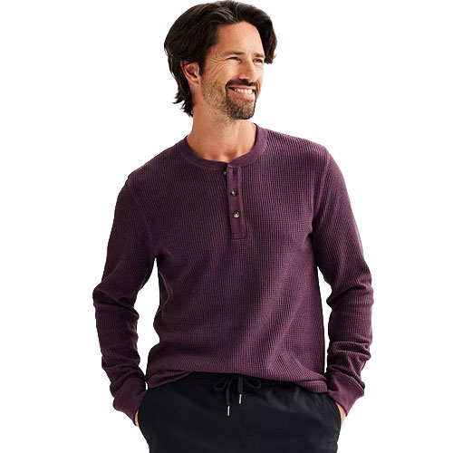 A man is wearing a long-sleeved, purple waffle-knit henley shirt with buttons and dark pants.