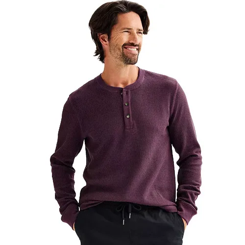 A man is wearing a long-sleeved, purple waffle-knit henley shirt with buttons and dark pants.
