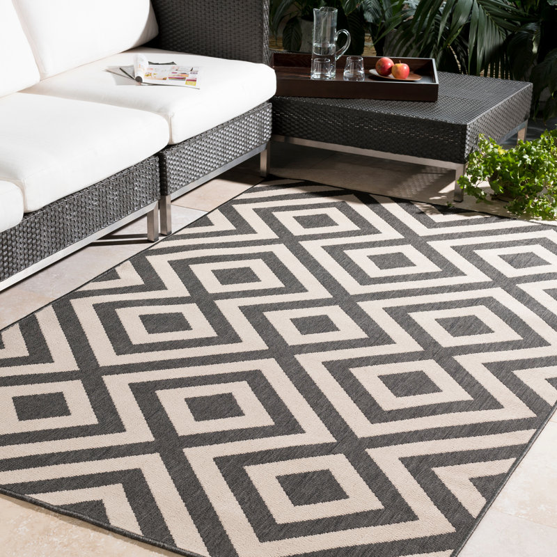 5' x 8' outdoor rug with a bold black and white geometric pattern featuring interconnected diamond shapes, placed on a patio near a rattan sofa and coffee table.