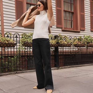 A person on a sidewalk wears a sleeveless ribbed turtleneck top, black wide-leg pants, and tan flat shoes, holding a tan coat and wearing sunglasses.