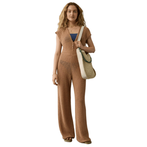 A woman is wearing a brown knit jumpsuit with wide legs and cap sleeves, carrying a woven shoulder bag with a chain detail. She accessorizes with layered necklaces and a bracelet.