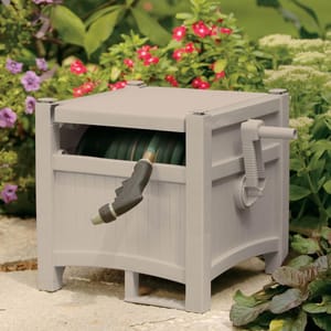 A garden hose reel box with a crank handle, storing a coiled hose with a nozzle, is placed among flowering plants.