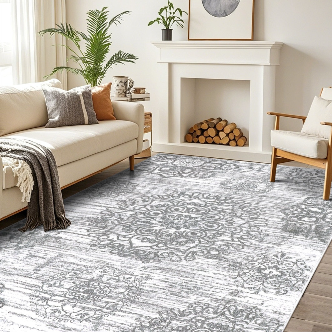 A modern living room features a light-colored sofa, an armchair, and a decorative rug with intricate patterns. A white fireplace is adorned with potted plants, and a stack of firewood is neatly arranged inside.