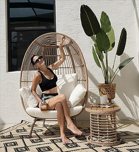 A round, cushioned egg chair made of an open-weave frame paired with a circular side table featuring a matching woven design, placed on a patterned rug outdoors.