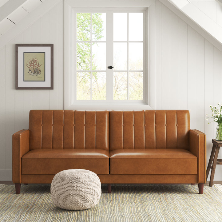 A tan leather sofa with tufted backrest, flanked by a wooden side table and a beige pouf, situated in front of a window with a view of trees.