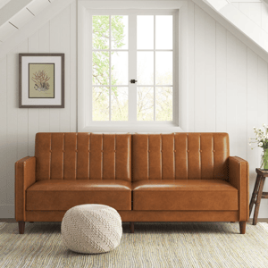 A tan leather sofa with tufted backrest, flanked by a wooden side table and a beige pouf, situated in front of a window with a view of trees.