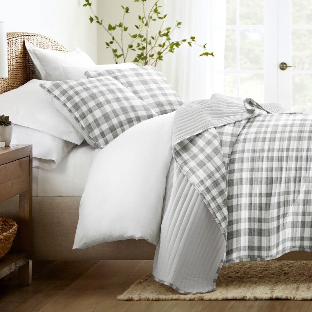 A neatly made bed features gray and white plaid pillows and a matching quilt, with white bedsheets. The setting includes a wooden side table and plants, creating a cozy bedroom atmosphere.