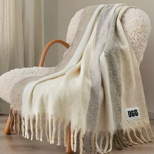 A cozy, striped UGG throw blanket with fringe detailing is draped over a textured armchair.
