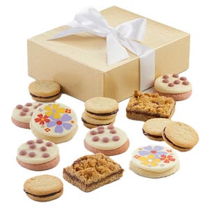 A gold box with a white ribbon contains an assortment of cookies, including round cookies with colorful floral designs, cookies with chocolate dots on white icing, sandwich cookies with filling, and crumb-topped square bars.