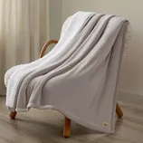 A soft gray blanket is draped over a cushioned wooden chair with curved wooden legs, set against a light wooden floor and neutral-toned walls.