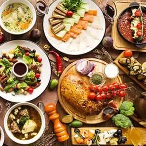A table filled with a variety of dishes including soups, salads, sliced meats, bread, and colorful vegetables, along with bowls of sauces and garnishes, showcasing a diverse and abundant meal spread.