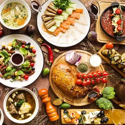 A table filled with a variety of dishes including soups, salads, sliced meats, bread, and colorful vegetables, along with bowls of sauces and garnishes, showcasing a diverse and abundant meal spread.