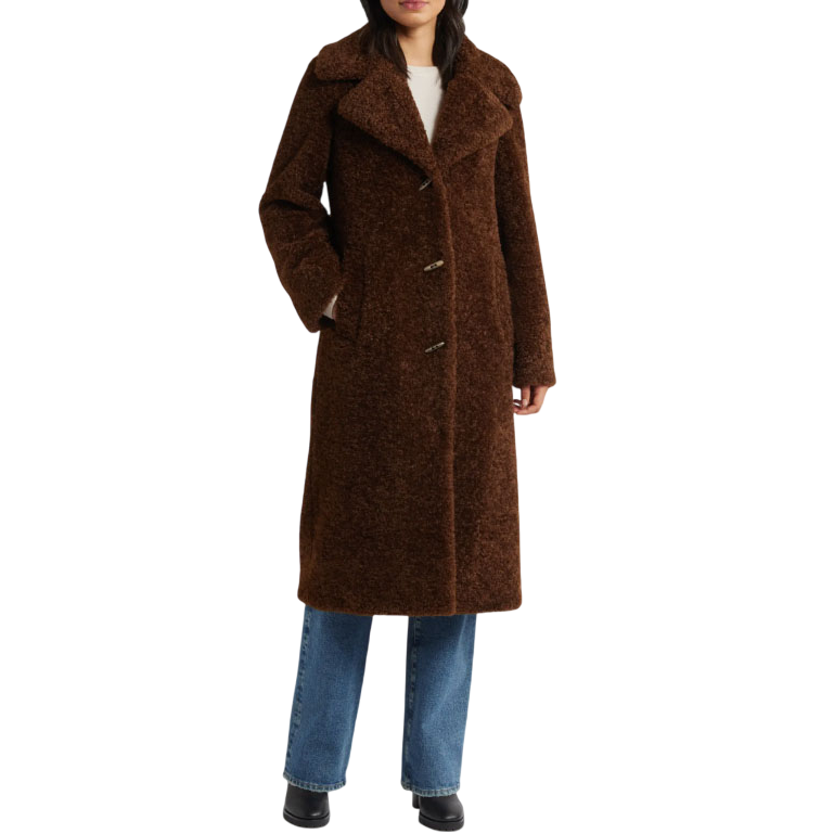 A woman is wearing a long, brown, textured coat with large lapels, over blue jeans and black shoes.