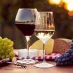 A glass of red wine and a glass of white wine, with grapes, cheese, and a corkscrew on a wooden table.