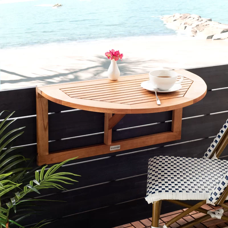 A half-round wooden balcony table with a ceramic vase and flowers on top, accompanied by a white cup and saucer, overlooking a beach view.
