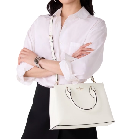 A woman is holding a white Kate Spade satchel with a top handle and an adjustable shoulder strap, featuring gold-tone hardware and a subtle logo on the front. She wears a white shirt and a black skirt.