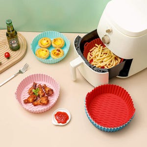 A white air fryer with fries inside, surrounded by silicone baking molds containing egg tarts and chicken wings, next to a bottle of beer.