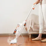 A person is using a white and orange steam mop on a wooden floor, releasing visible steam, with a sofa in the background. The mop is corded and suitable for cleaning hard surfaces.