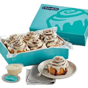 A box of six Cinnabon cinnamon rolls with icing, displayed alongside an extra container of frosting and one roll served on a plate with a fork.