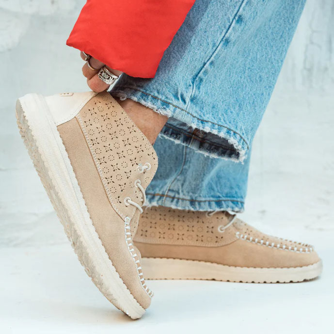 Beige suede boots with perforated detailing paired with blue distressed jeans and a glimpse of a red jacket.