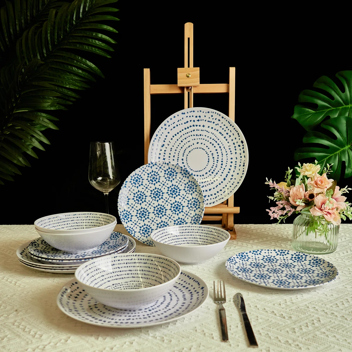 A dinnerware set featuring plates and bowls with blue patterns on a white background, alongside a wine glass, cutlery, and a vase with flowers, arranged on a textured tablecloth with leafy plants in the background.
