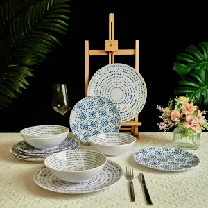 A dinnerware set featuring plates and bowls with blue patterns on a white background, alongside a wine glass, cutlery, and a vase with flowers, arranged on a textured tablecloth with leafy plants in the background.