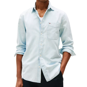 A person is wearing a light blue denim shirt with a chest pocket, rolled-up sleeves, and partially buttoned over a white undershirt, paired with dark pants.