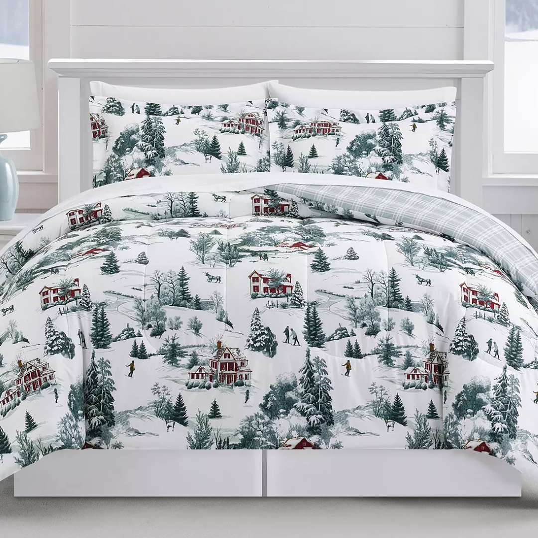 A winter-themed bedding set featuring a snowy village scene with red houses and evergreen trees. The comforter is reversible with a plaid pattern on the other side.