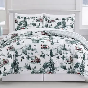 A winter-themed bedding set featuring a snowy village scene with red houses and evergreen trees. The comforter is reversible with a plaid pattern on the other side.