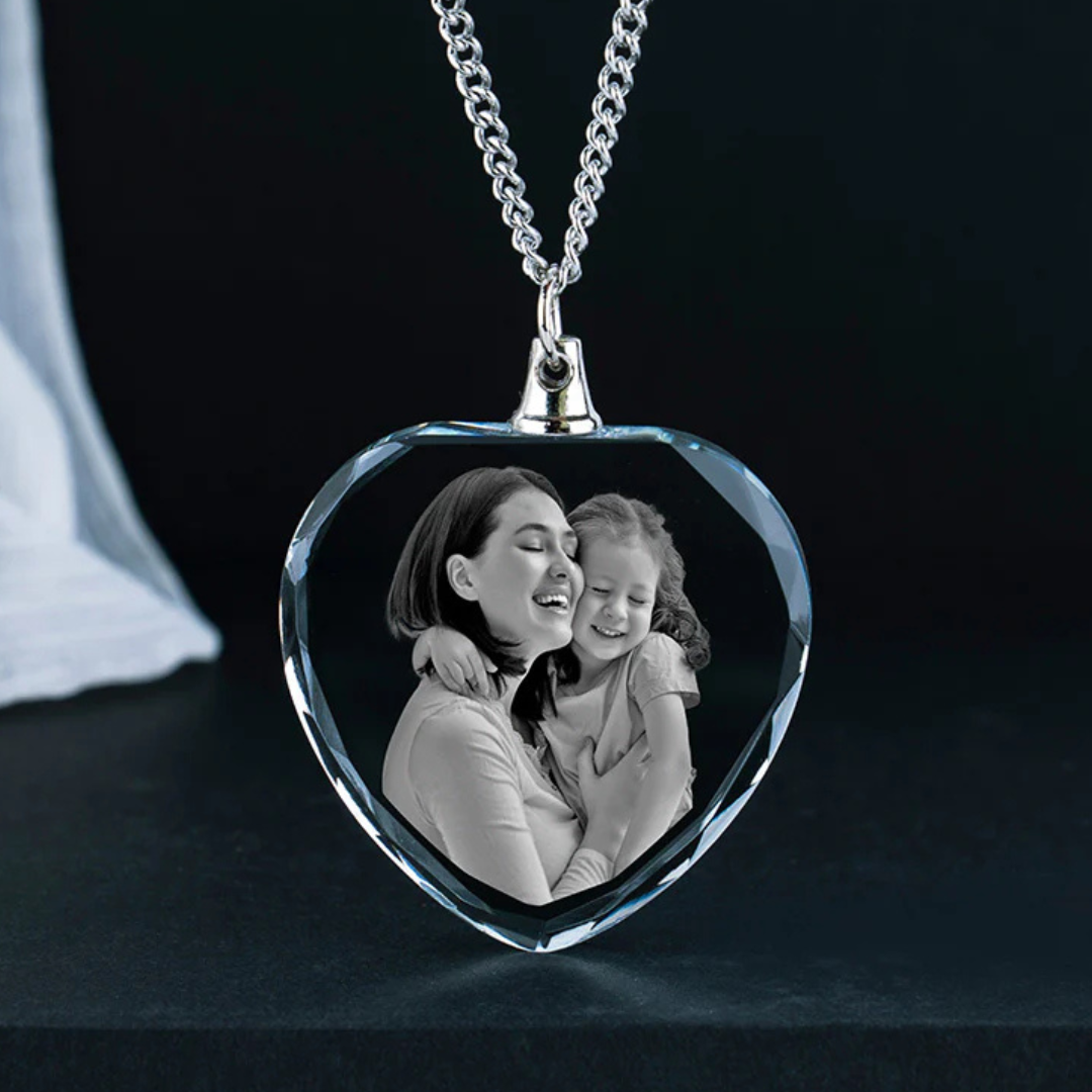 A heart-shaped crystal pendant necklace features a black-and-white photo of a woman and child embracing, with the image elegantly engraved inside the crystal.