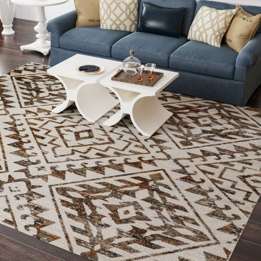 A modern living room setup featuring a geometric-patterned area rug on a wooden floor, with a blue sofa adorned with patterned cushions and two white nesting tables holding glasses and a serving tray.