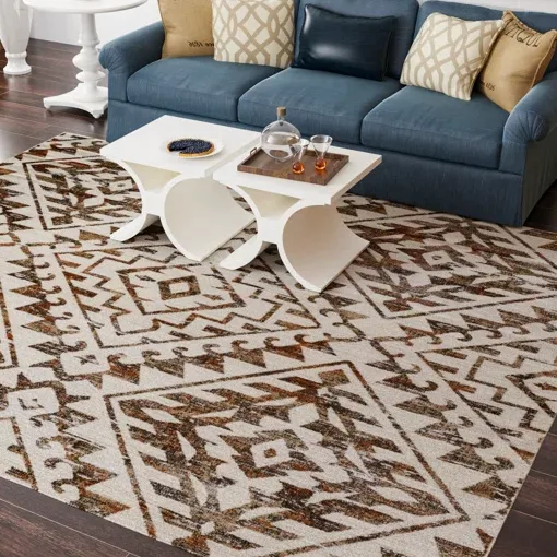 A modern living room setup featuring a geometric-patterned area rug on a wooden floor, with a blue sofa adorned with patterned cushions and two white nesting tables holding glasses and a serving tray.