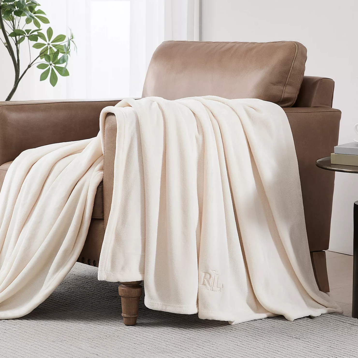 A soft, cream-colored throw with \“RL\“ initials draped over a tan leather armchair, alongside a small round side table holding a cup and books, set in a minimalistic room with white curtains and a potted plant.