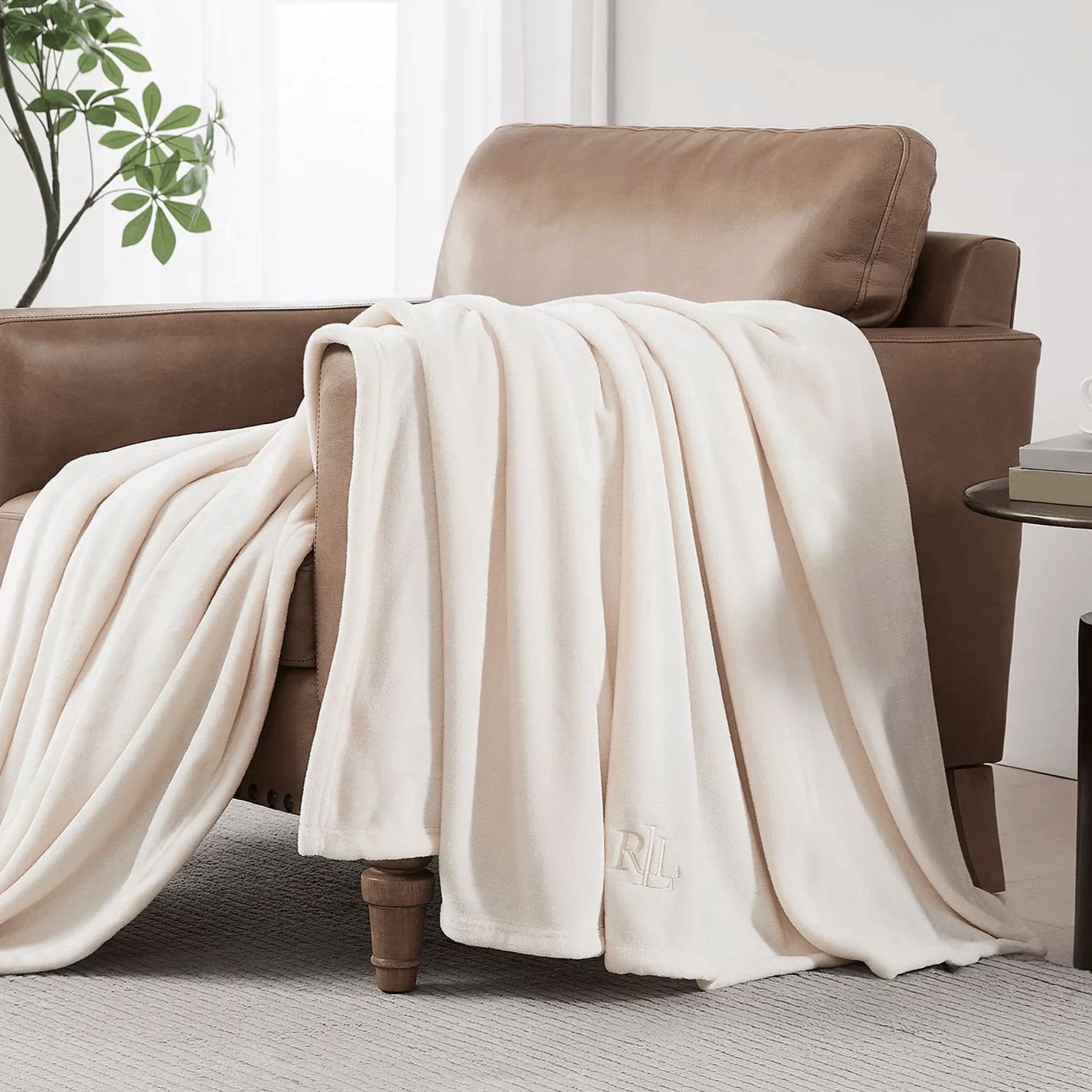 A soft, cream-colored throw with \“RL\“ initials draped over a tan leather armchair, alongside a small round side table holding a cup and books, set in a minimalistic room with white curtains and a potted plant.