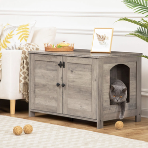 A gray wooden cabinet with dual doors and a side cutout functions as a pet house, with a tray and framed artwork on top. A gray cat is partially inside and outside the cabinet, with yarn balls nearby.